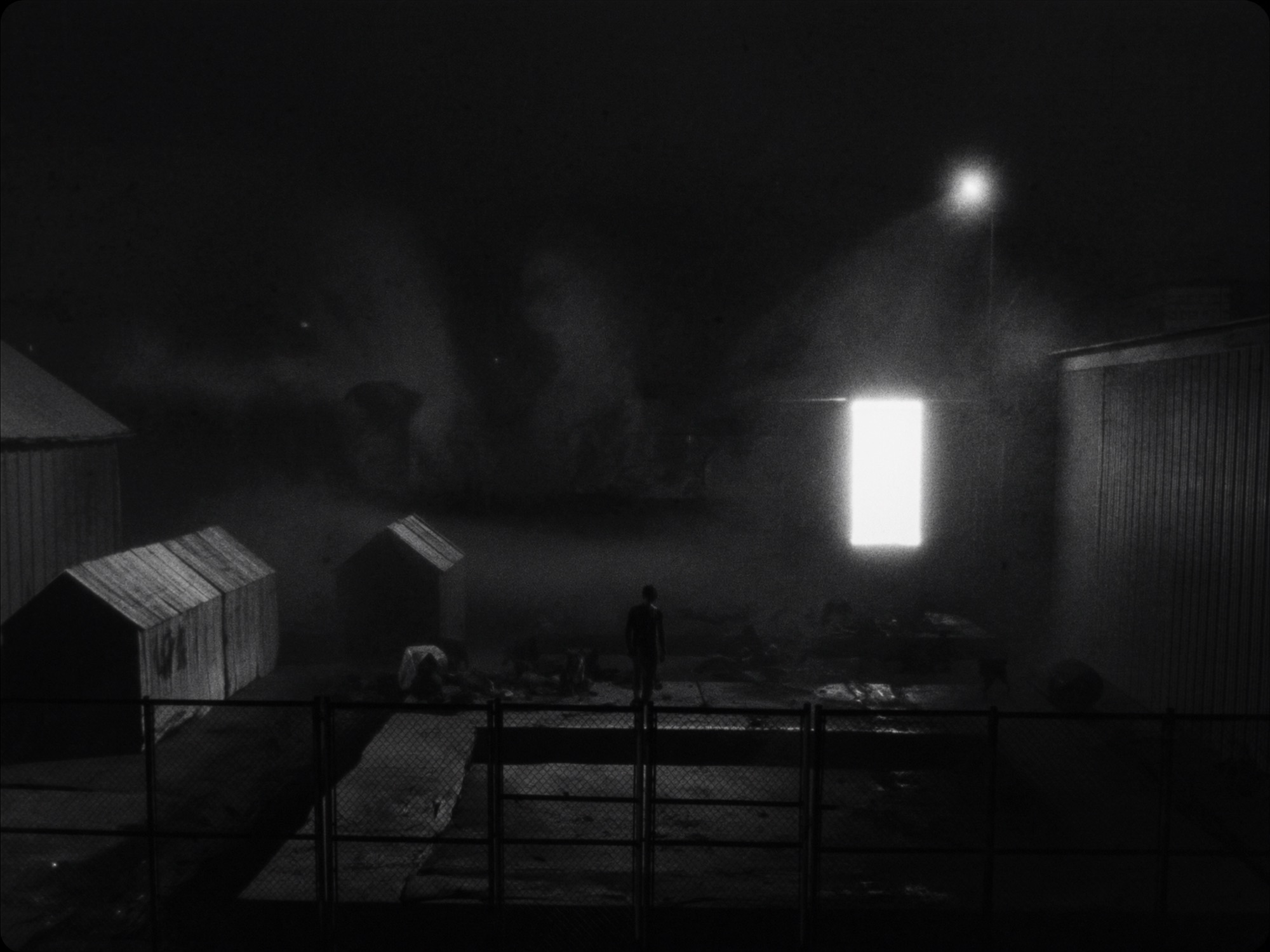 A lit doorway in a foggy yard at night, a figure walking toward it